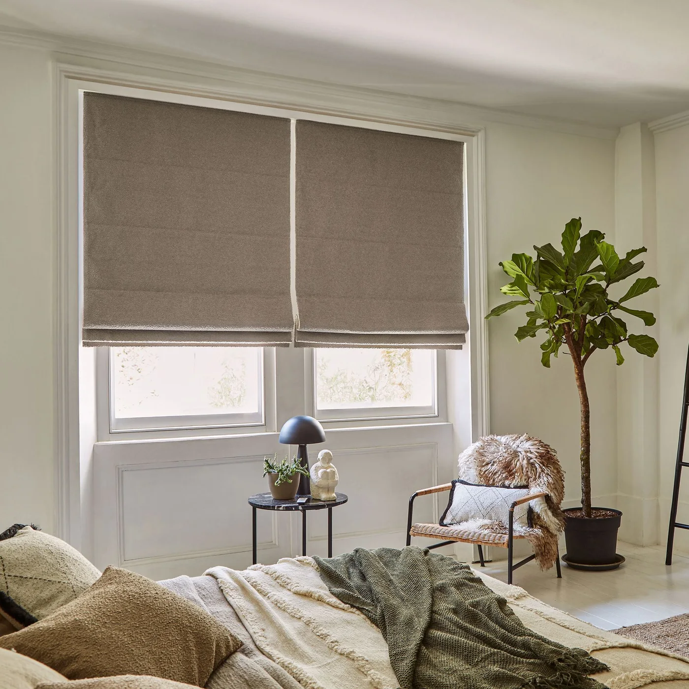 A textured chair and small dark side table sit beneath broad taupe blinds beside a leafy plant and a bed with pale and green layers