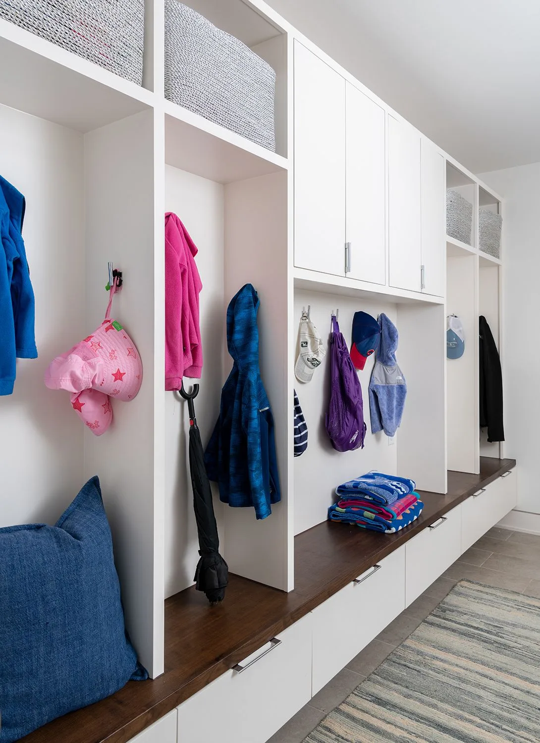 White mudroom with divided hanging areas, a long wood bench, upper baskets and closed cupboards in the center