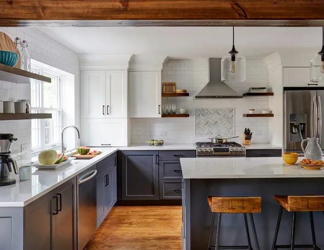 Dark blue lower cabinets and an island sit beneath white upper cupboards and an exposed wooden beam beside a metallic hood and warm wooden stools