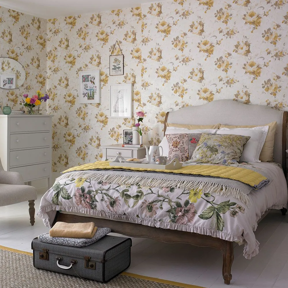 Bedroom with floral wallpaper, a wooden bed, botanical bedding, pale drawers, framed pictures and a small suitcase at the foot.