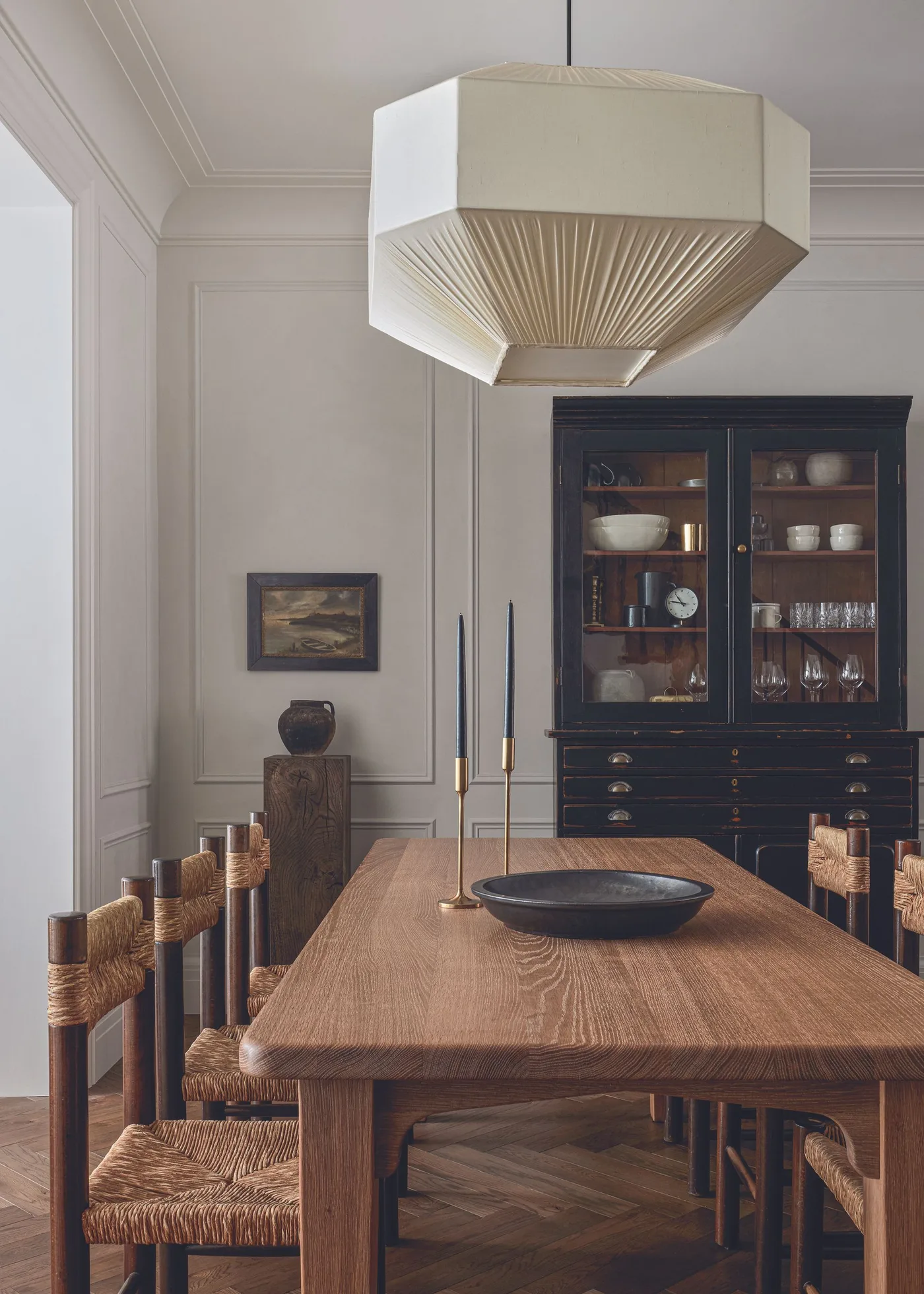 Large pale geometric pendant with a pleated underside above a wood dining table and woven-seat chairs