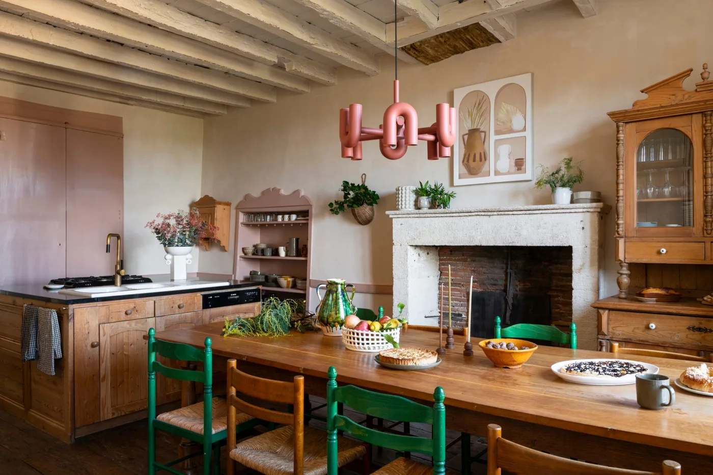Wooden table with green and yellow chairs beside a fireplace, a tall display cabinet and a pink ceiling light.