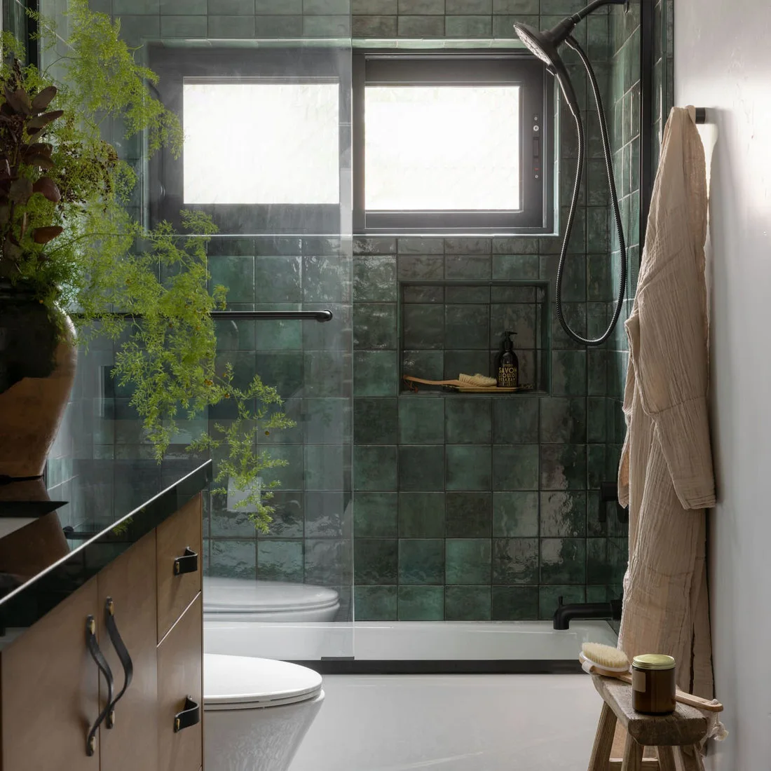 Deep green rectangular shower tiles surround a window and niche beside a wood-fronted vanity and pale walls