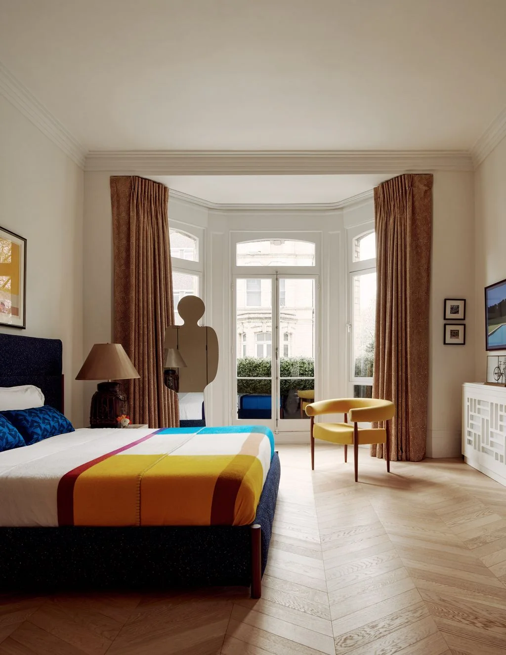 A bed with dark headboard and a colorful geometric throw stands beside warm curtains, a yellow open-backed chair and pale walls near a bay window