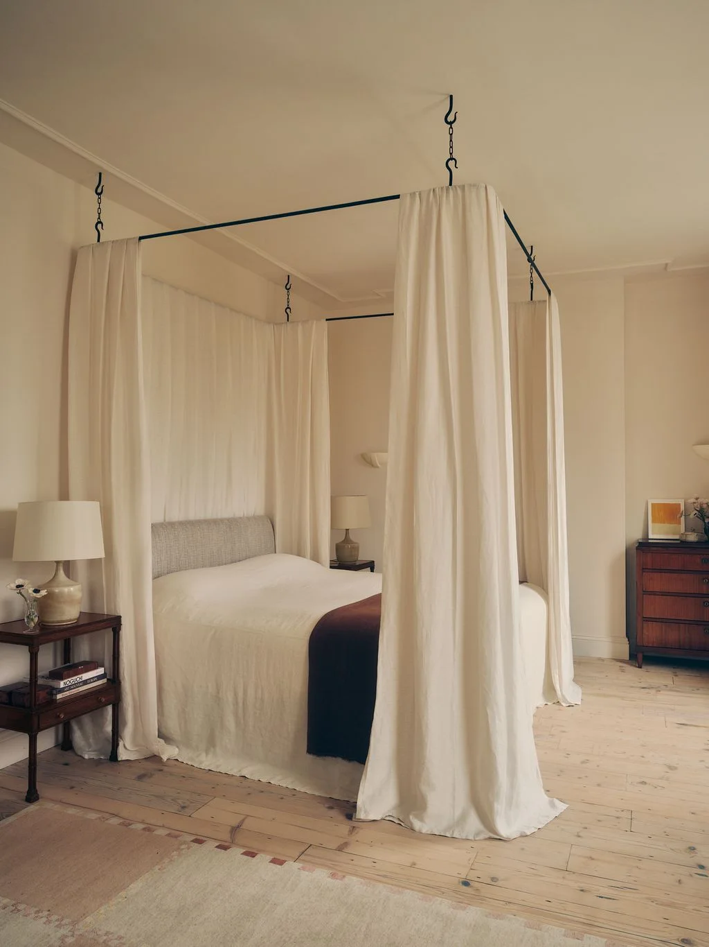 A pale bed stands beneath a thin suspended dark canopy frame with long light curtains beside small wood bedside tables and pale walls
