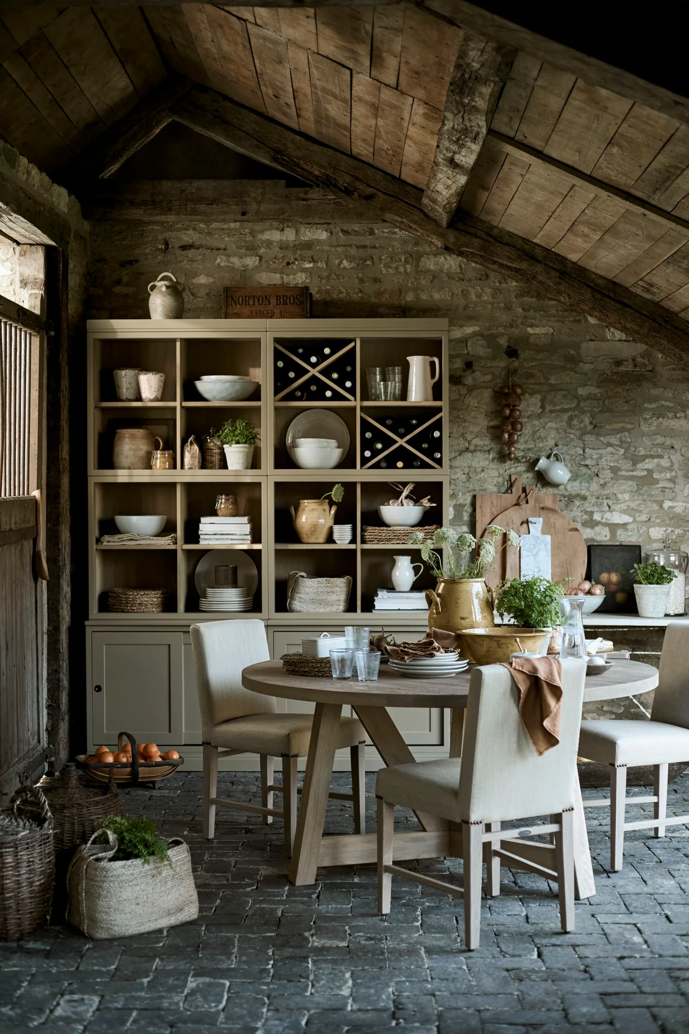 Pale dining hutch with open compartments, bottle racks and lower cupboards beside a wood table and white chairs