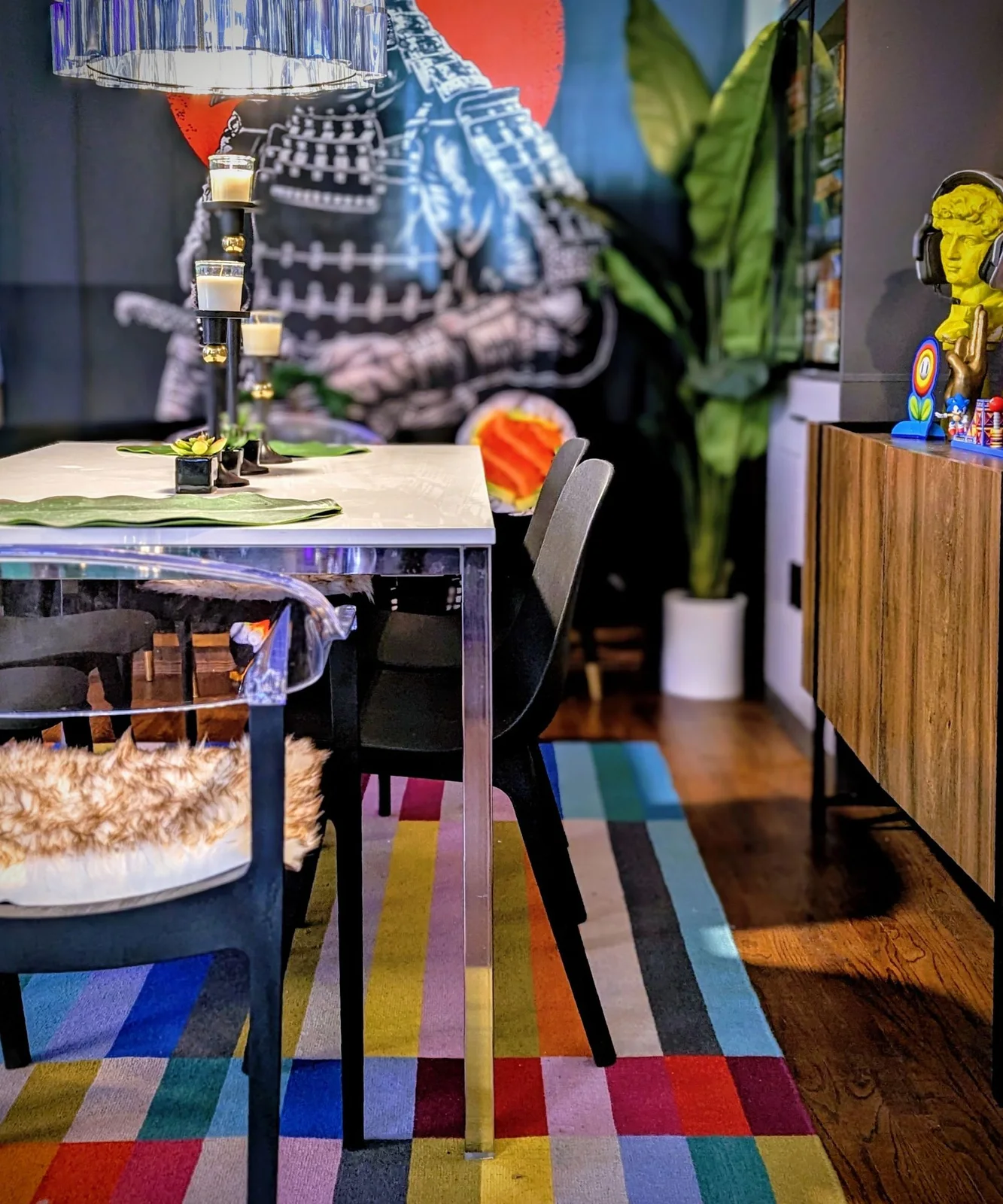 A wood-fronted sideboard with dark legs stands beside a reflective dining table, dark chairs and a multicolored striped rug