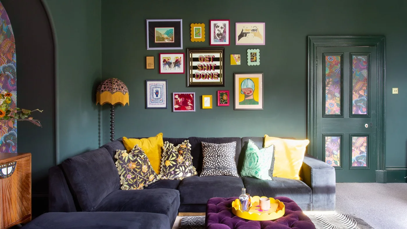 Deep green walls surround a dark corner sofa beneath a colorful gallery of framed pictures