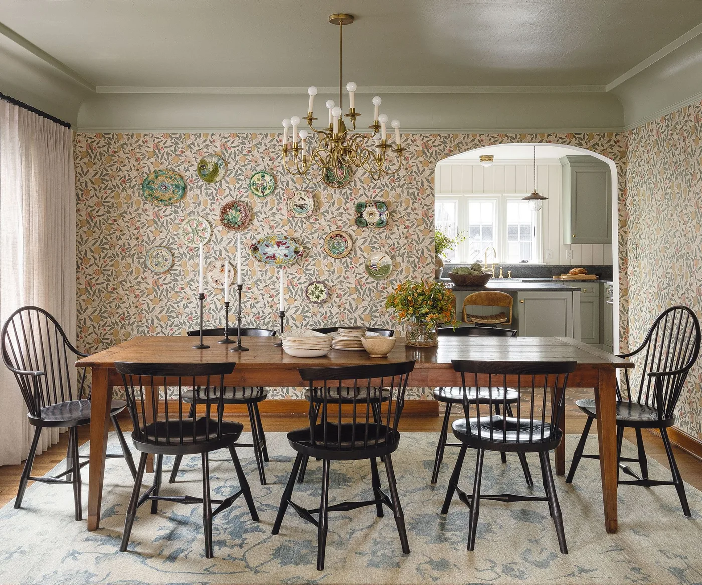 Cottage dining room with small floral wallpaper, black spindle-back chairs, wooden table, plate display and an opening toward a pale kitchen.