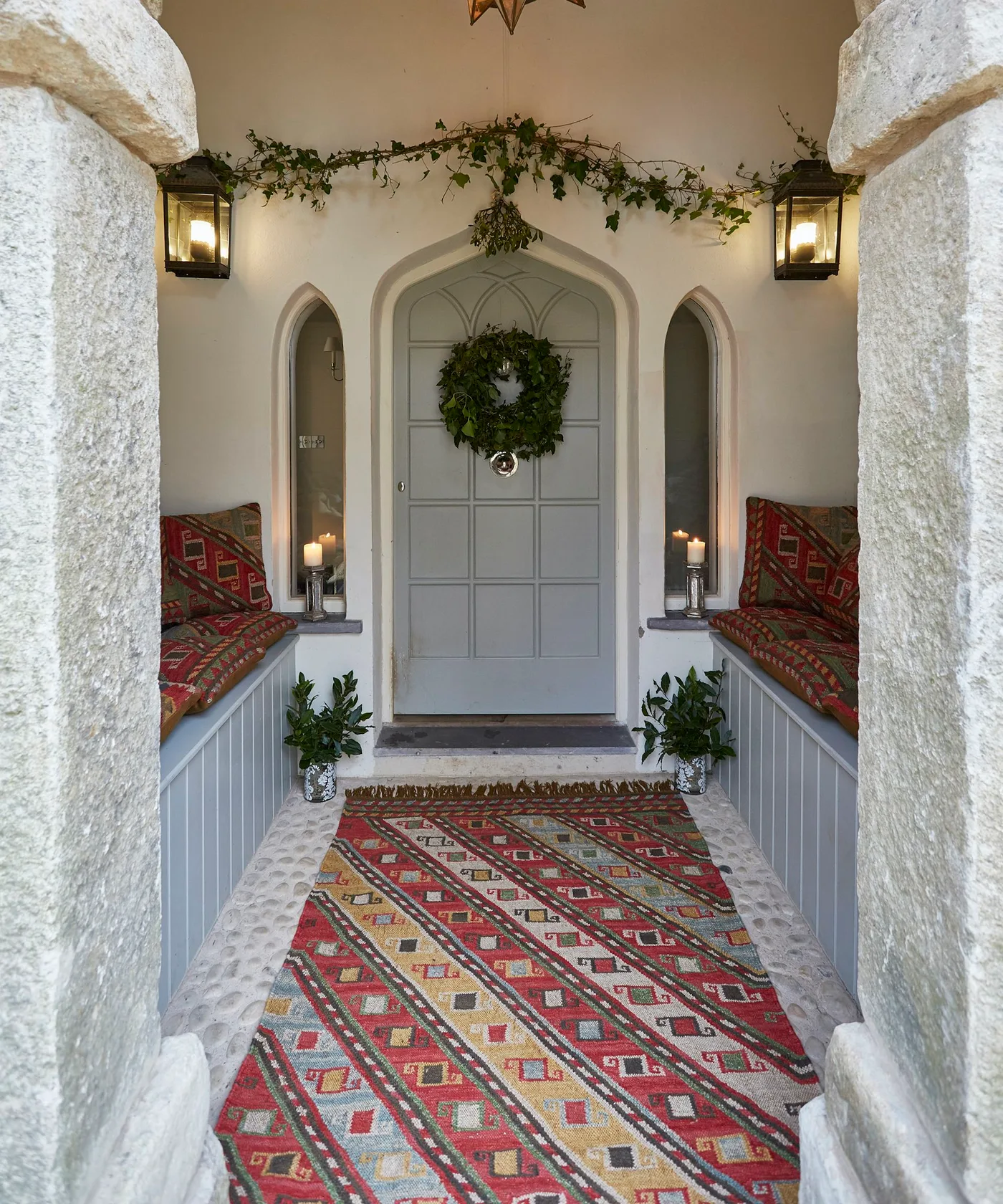 Red and yellow geometric rug between cushioned built-in porch benches, pale paneled door, plants and rough stone doorway