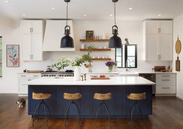 A navy island stands below two dark pendants in a pale kitchen with wooden stool seats, warm open shelves and a bright sink window