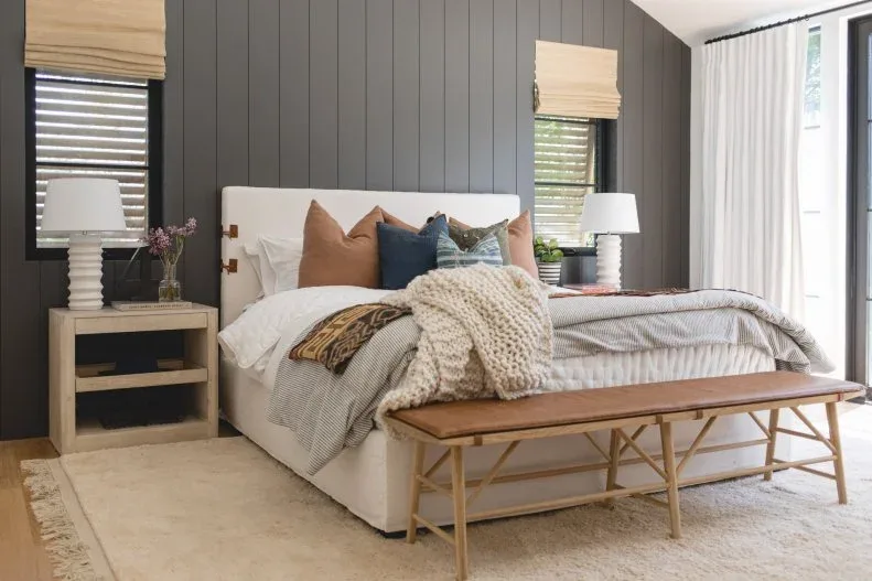 Bedroom with charcoal vertical wall boards, pale upholstered headboard, layered bedding and foot bench