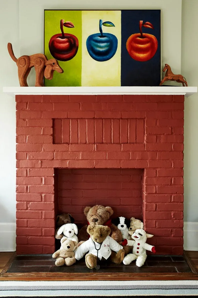 Red brick fireplace with a white mantel, colorful apple painting and stuffed toys in its opening.