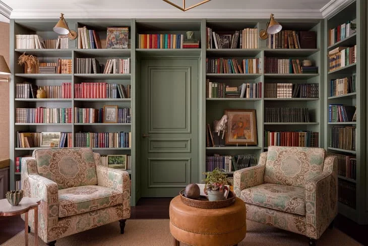 Green bookcases surround a matching central door behind two patterned chairs and a round brown ottoman