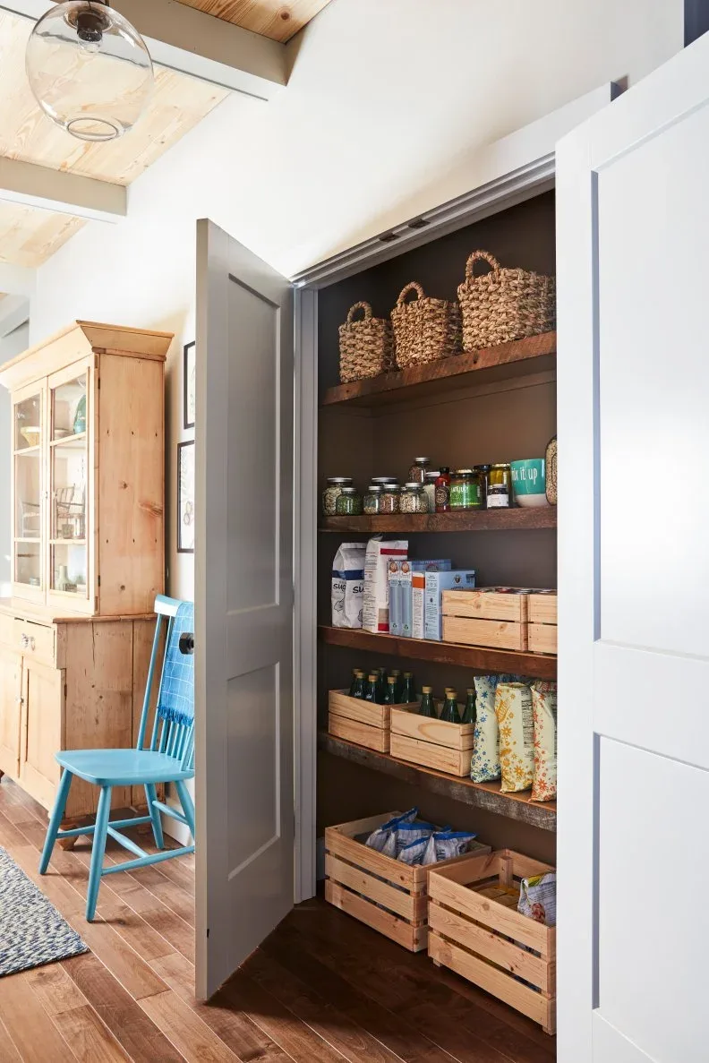 Pantry behind pale doors with dark background, wood shelves and crates, glass jars and woven baskets
