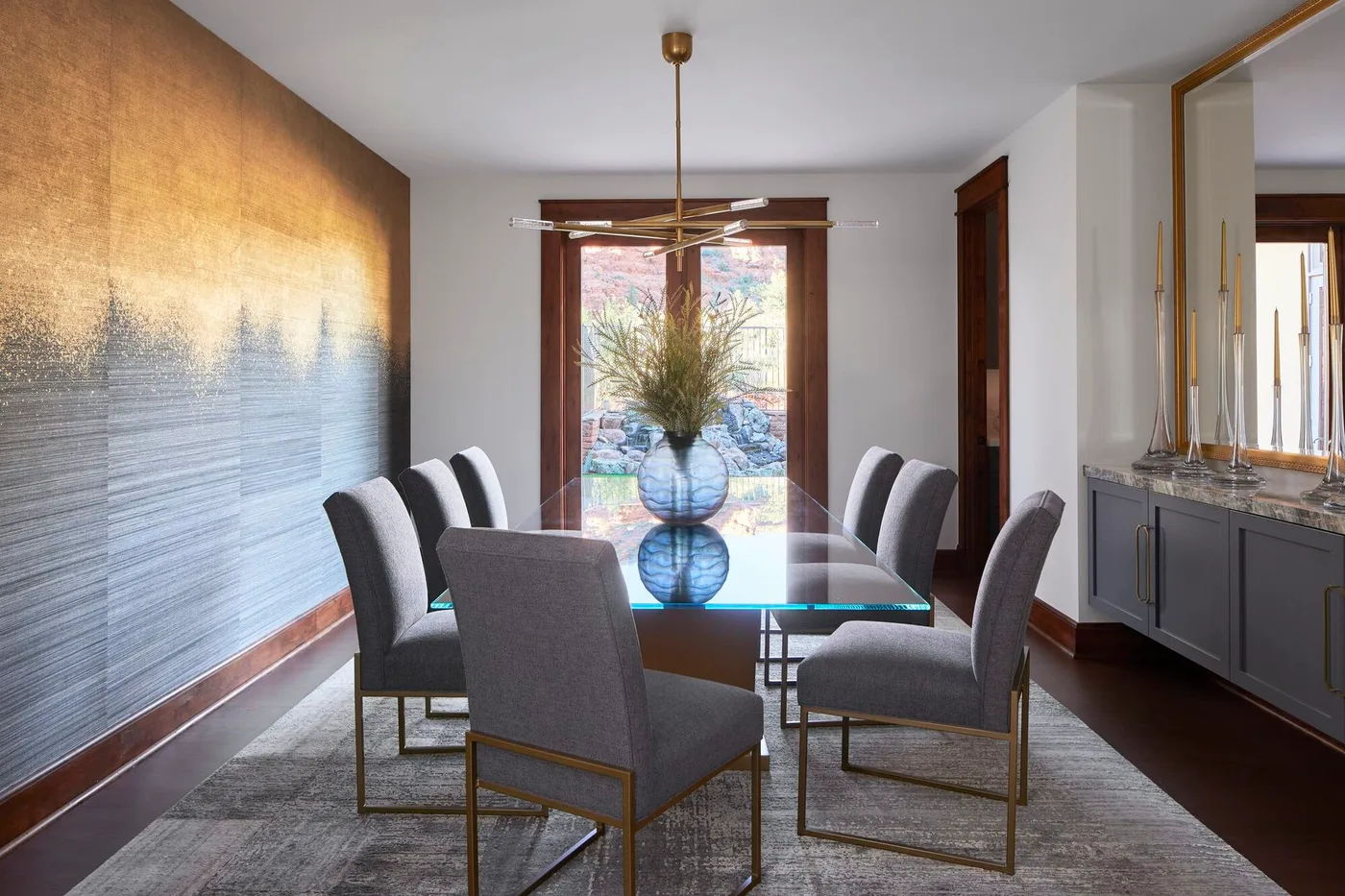 Clear rectangular glass dining table with gray upholstered chairs, a textured rug and warm wood window trim