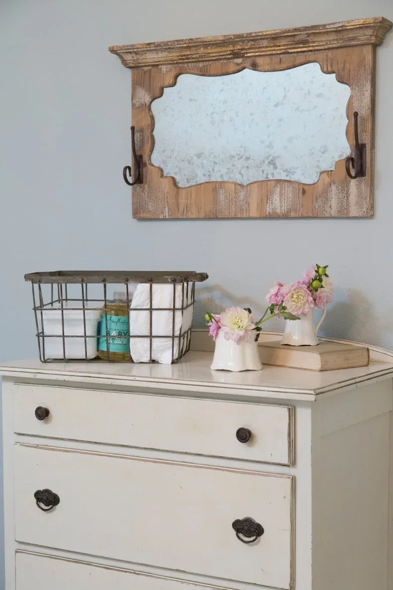 Pale dresser beneath decorative wooden mirror on blue-gray wall with small flower vessels and wire basket.