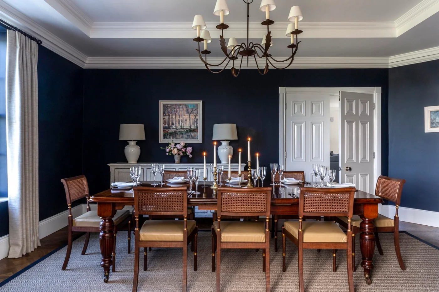 Navy walls surround a warm wooden dining table and woven-backed chairs beneath a pale ceiling with a chandelier, light curtains and white double doors