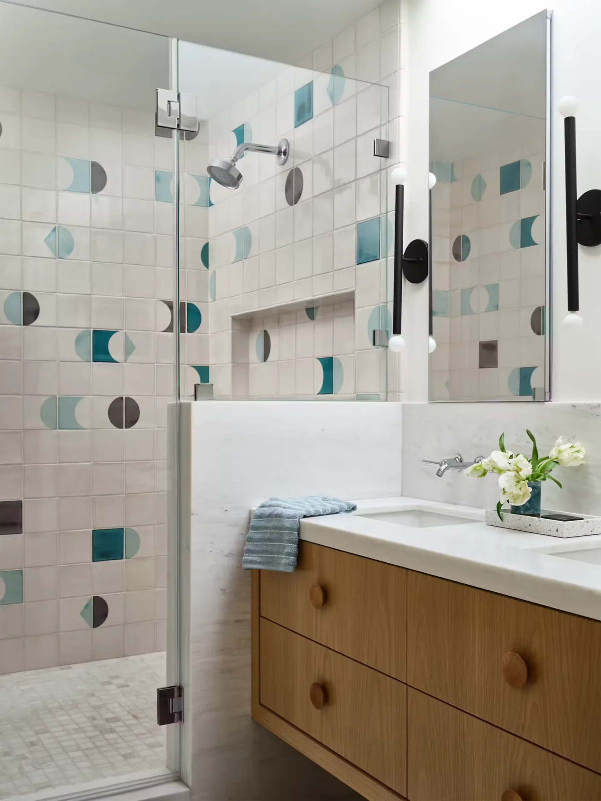 Pale bathroom tiles with scattered blue and gray geometric shapes surround a glass shower and mirrored cabinet above a wood vanity with round handles