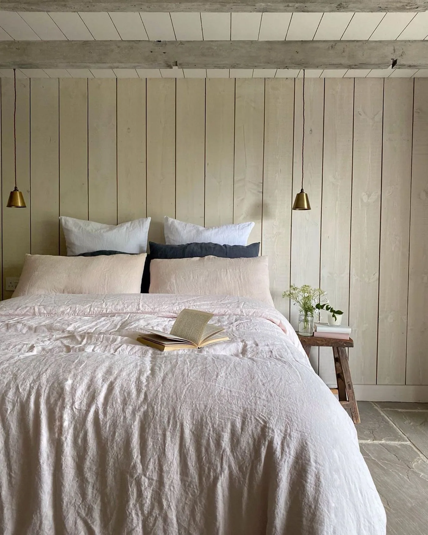 Country bedroom with pale vertical wall boards, a white bed, dark narrow pillows, two small hanging lights and a wooden bedside stool.