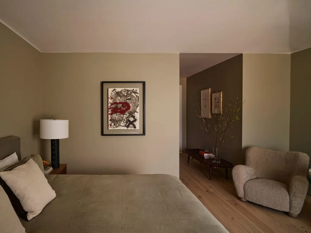 A pale bed and beige upholstered chair sit beneath simple artwork against warm muted walls beside a small bedside lamp and light wood flooring