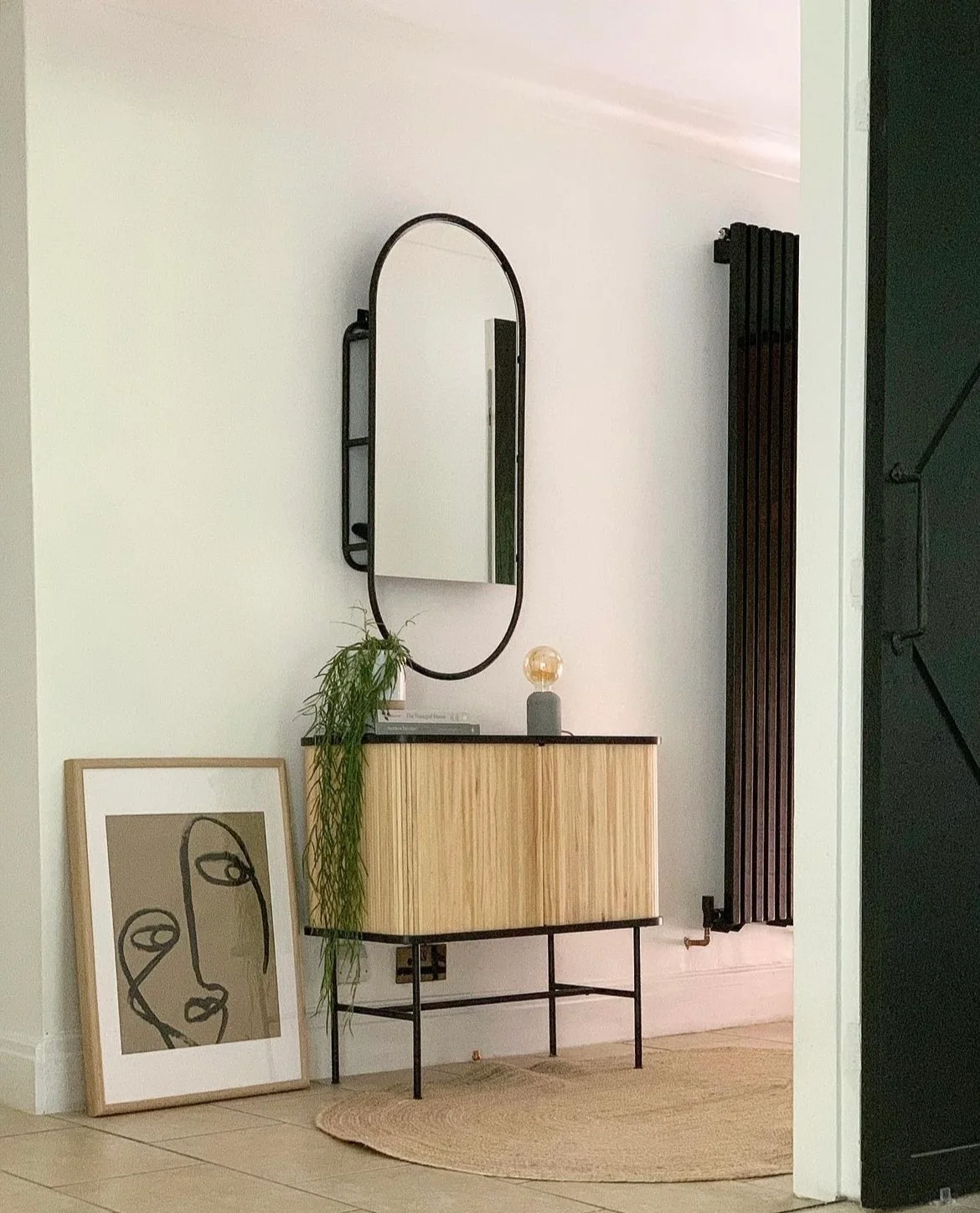 A reeded wooden cabinet with dark legs stands beneath an oval mirror beside a black radiator, trailing plant and round woven rug