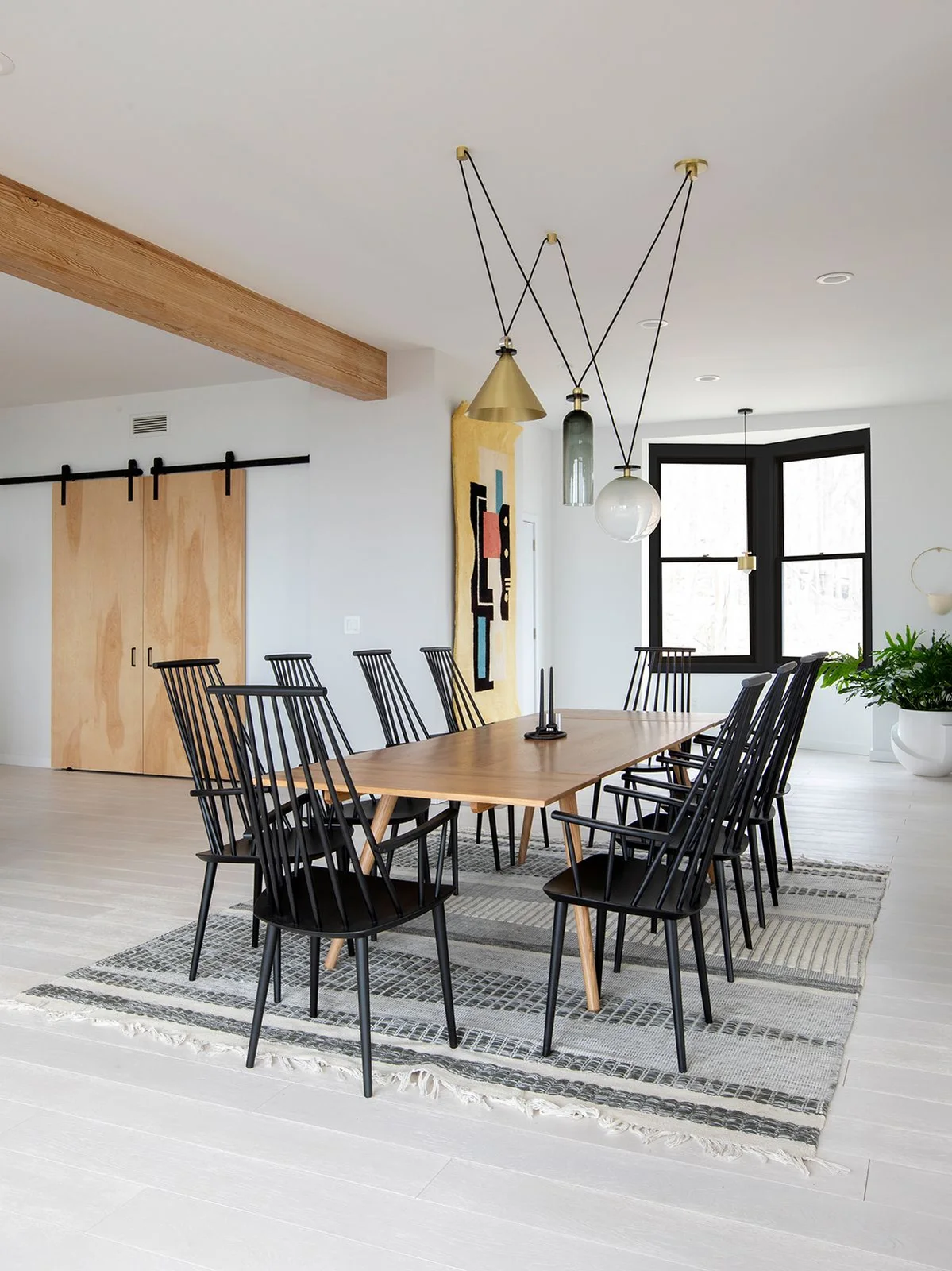 Black spindle-back dining chairs surround a pale wood table on a striped neutral rug below mixed hanging lights beside white walls and dark window trim