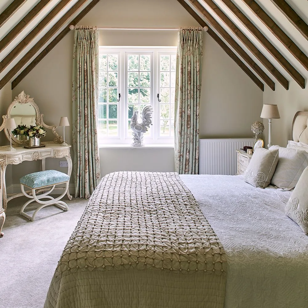 Bedroom with exposed sloping beams, pale walls, patterned curtains, layered pale bedding and a small decorative dressing table.
