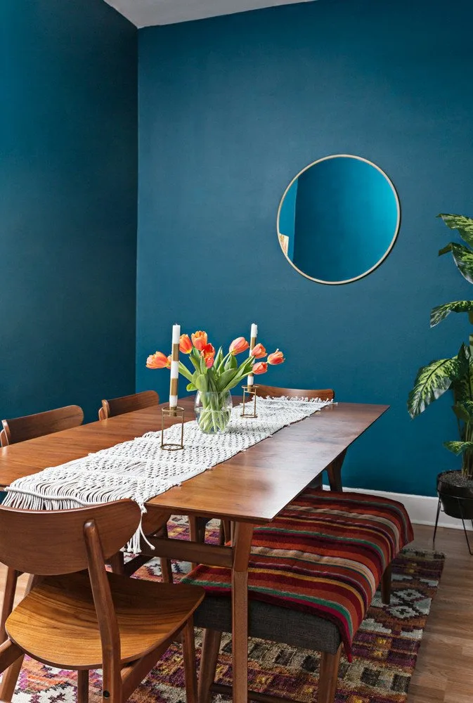 Round thin-framed mirror on a deep blue wall behind a wood dining table and striped bench