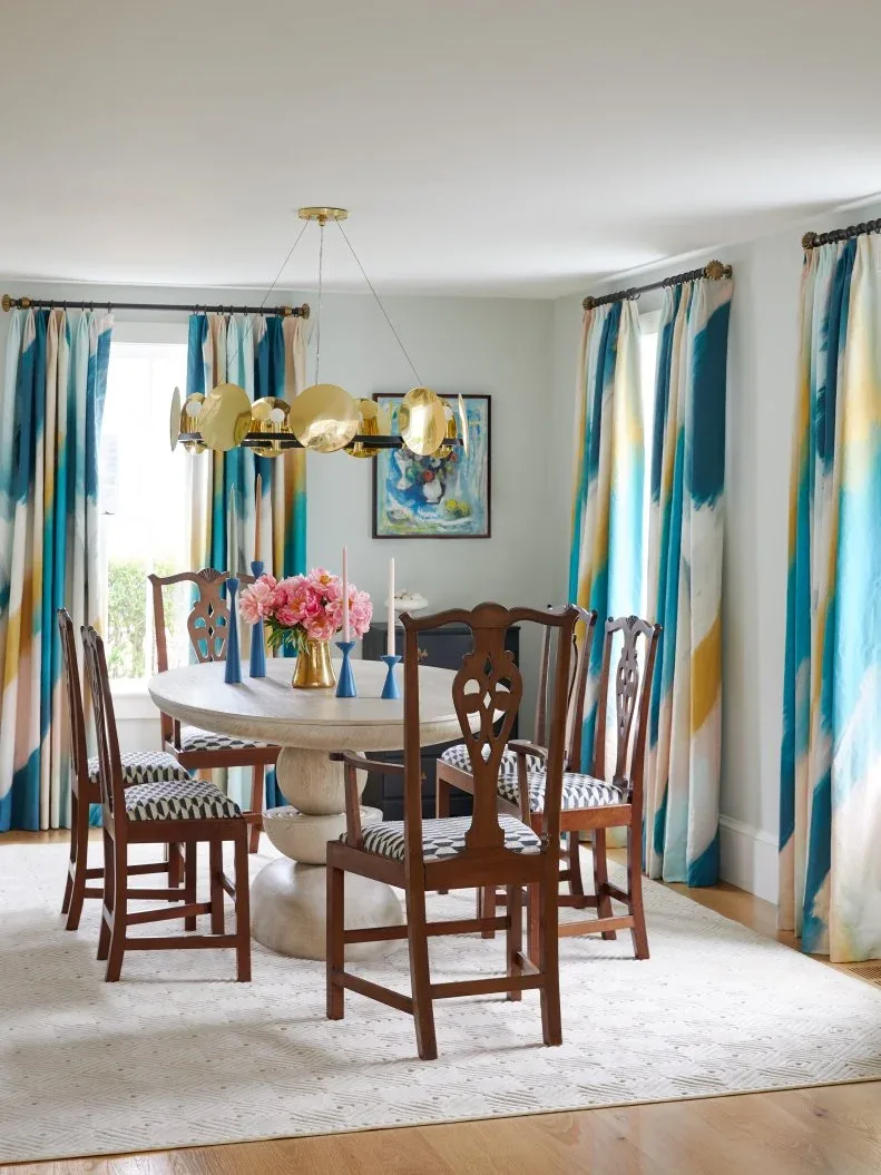Blue, yellow, and white abstract picture between windows with coordinating patterned curtains, above a pale round table, carved chairs, and a gold-colored circular chandelier.