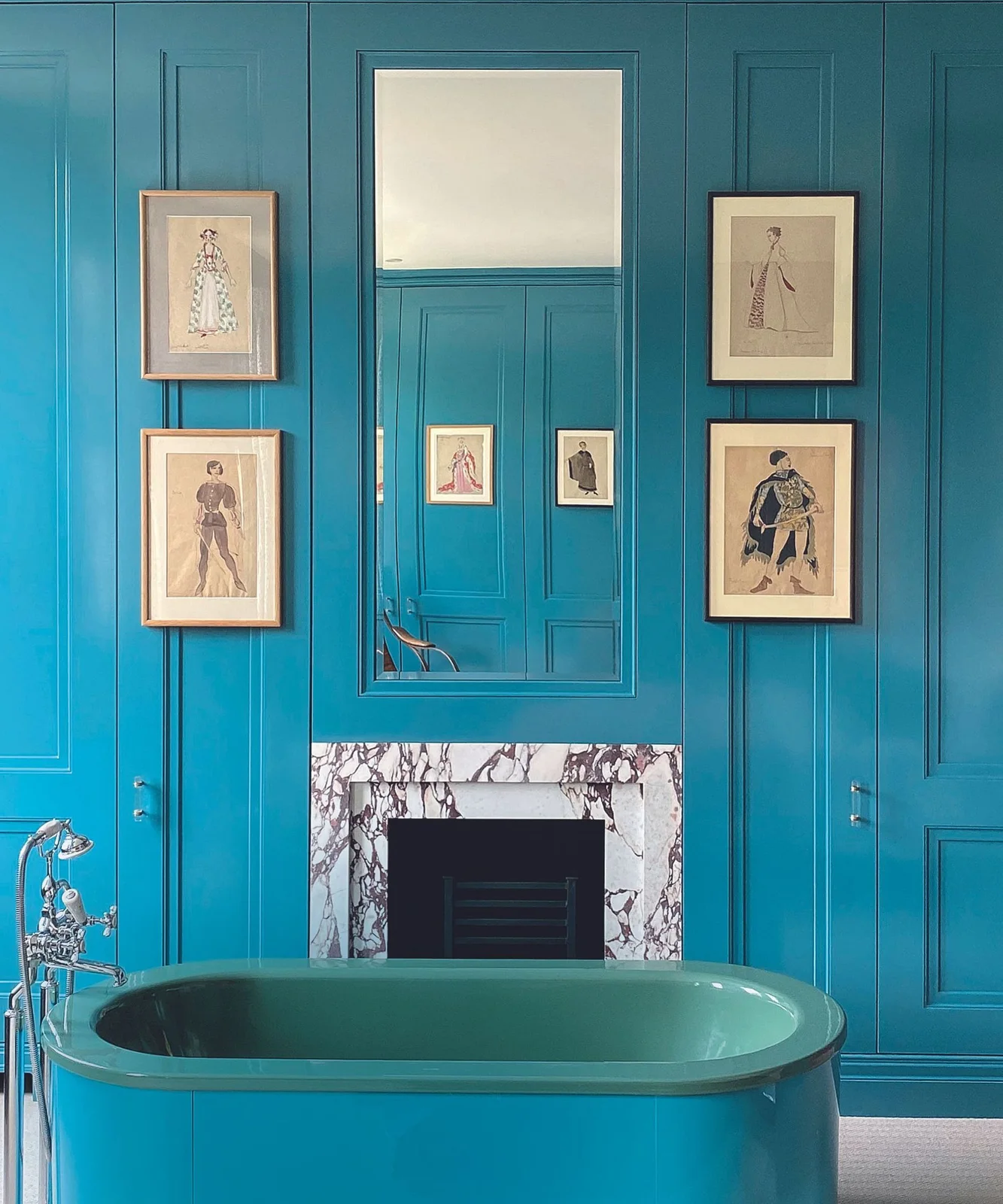 A blue freestanding bath stands before blue paneled walls with framed fashion pictures and a veined fireplace surround