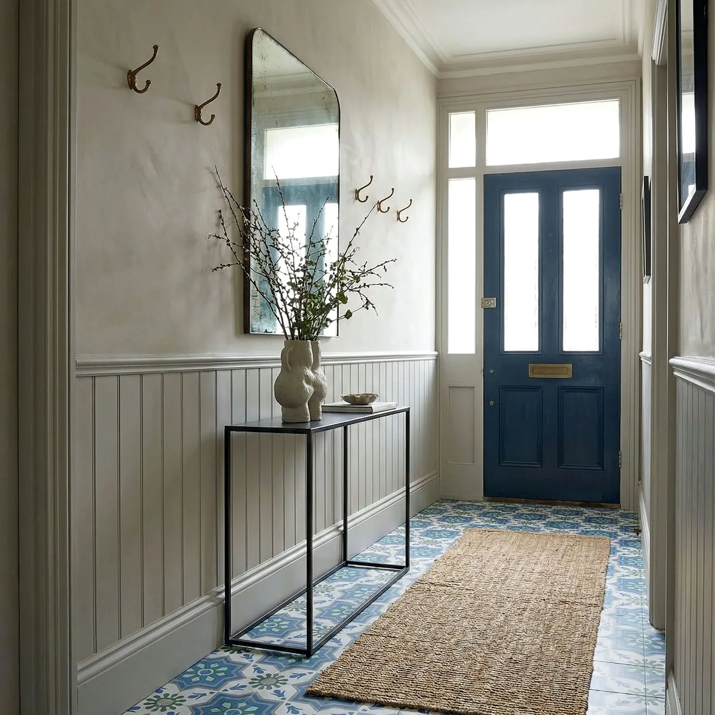 Pale paneled entry with dark blue glazed door, tall shaped mirror, wall hooks, thin black console, patterned floor and plain rug