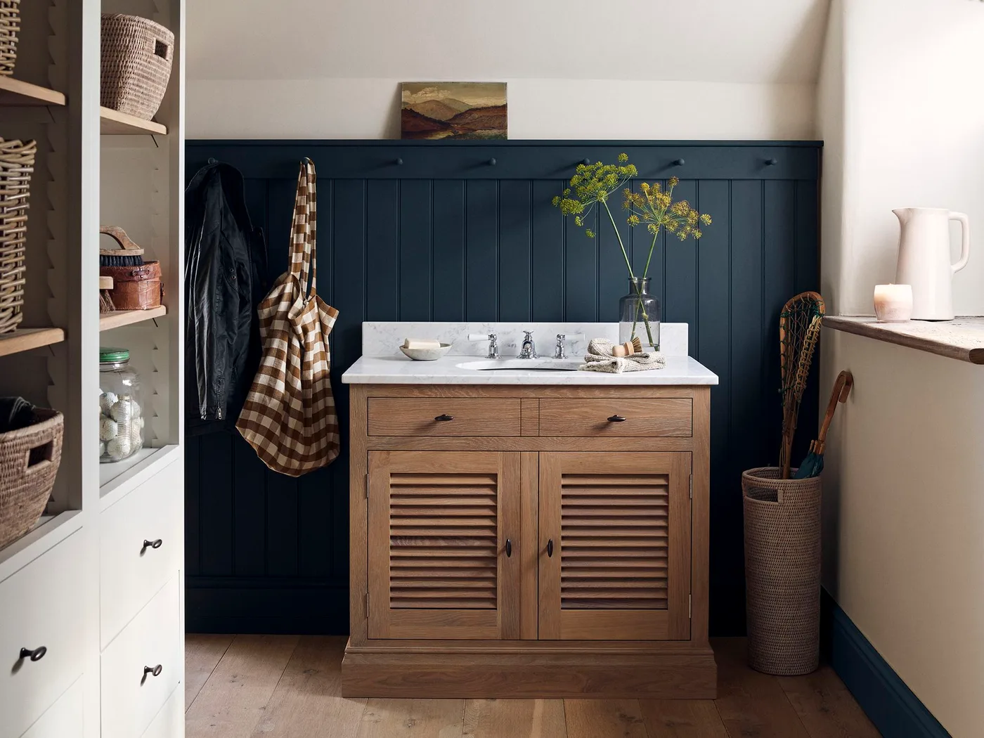 Country cloakroom with navy paneling, wooden louvered-door washstand, pale countertop and shelves holding baskets and jars.