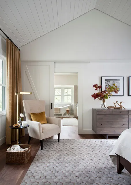 A light bedroom includes an upholstered chair near the window and timber storage furniture.