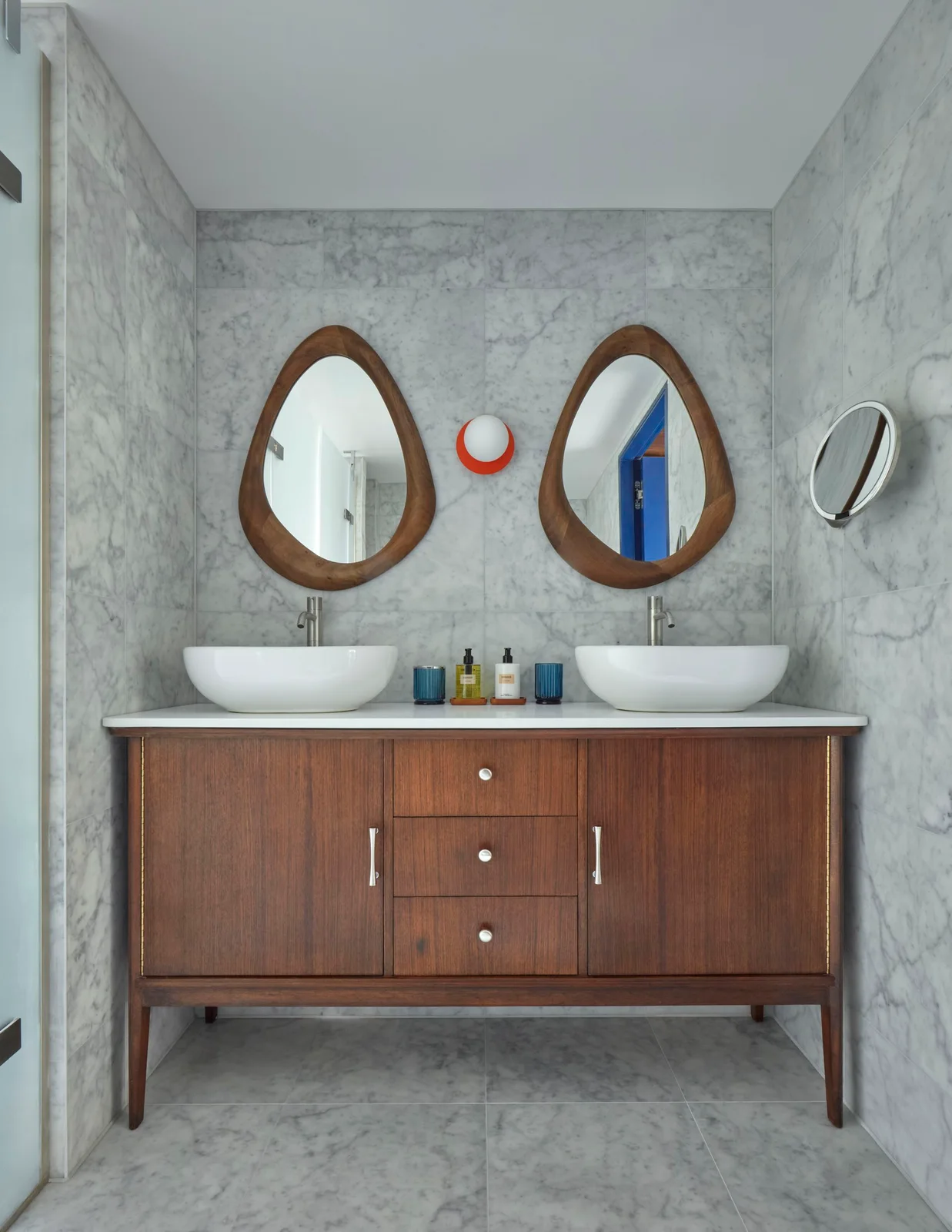 A dark wood bathroom vanity with tapered legs supports two white basins beneath curved wood-framed mirrors beside pale veined walls and a small red globe light