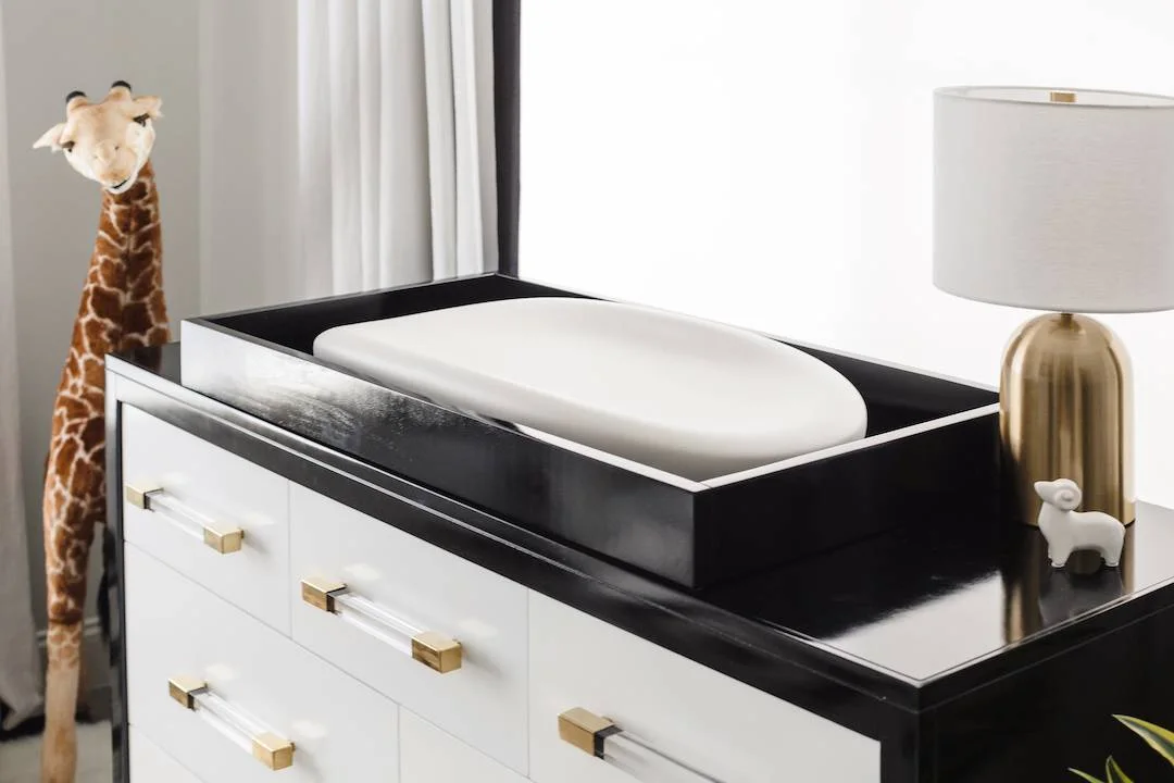 A nursery drawer chest has white fronts within a dark frame beneath a pale changing pad and white-shaded lamp beside light curtains