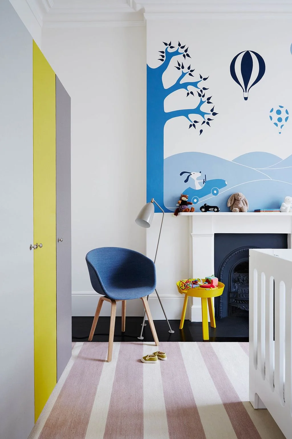 A blue illustrated nursery mural meets a pale striped rug beside a blue adult chair, yellow table and yellow-and-gray cupboard doors