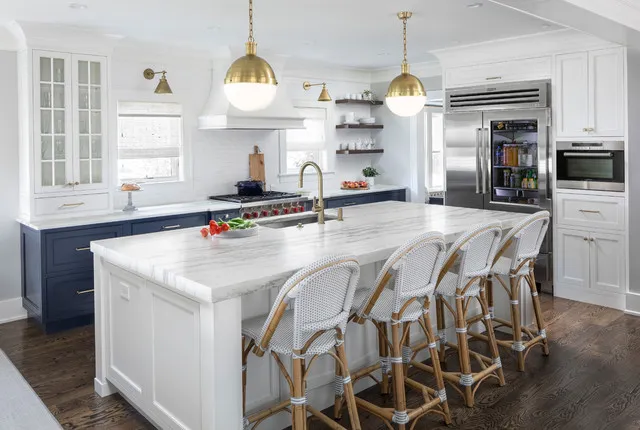 Navy lower cabinets surround a pale island with woven-backed chairs beneath rounded warm-colored pendants beside white upper cupboards and appliances