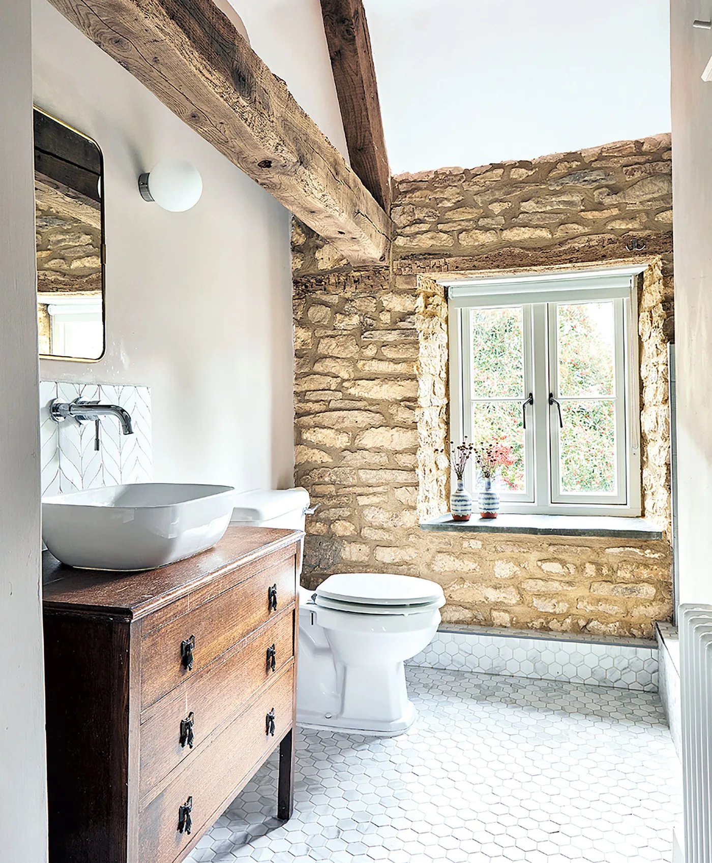 Bathroom with an exposed stone wall and beams, wooden drawer washstand, white vessel basin and pale small hexagonal floor tiles.