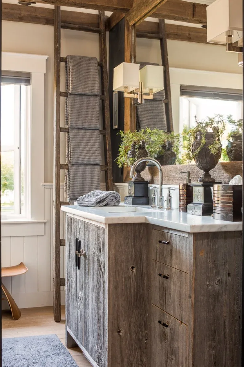 A rustic wood vanity with drawers and a pale top stands beside a large mirror, hanging square shades and a ladder holding folded towels