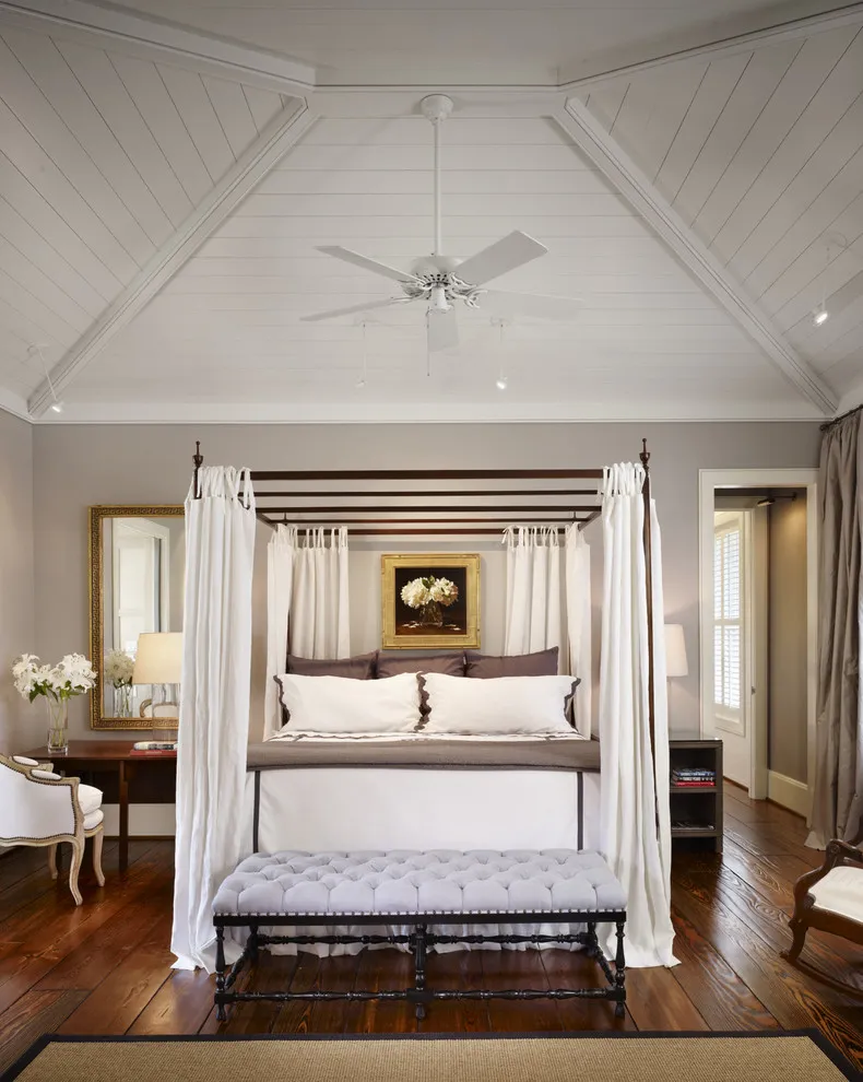 A dark canopy bed with white curtains stands on brown timber flooring beneath a pale roof