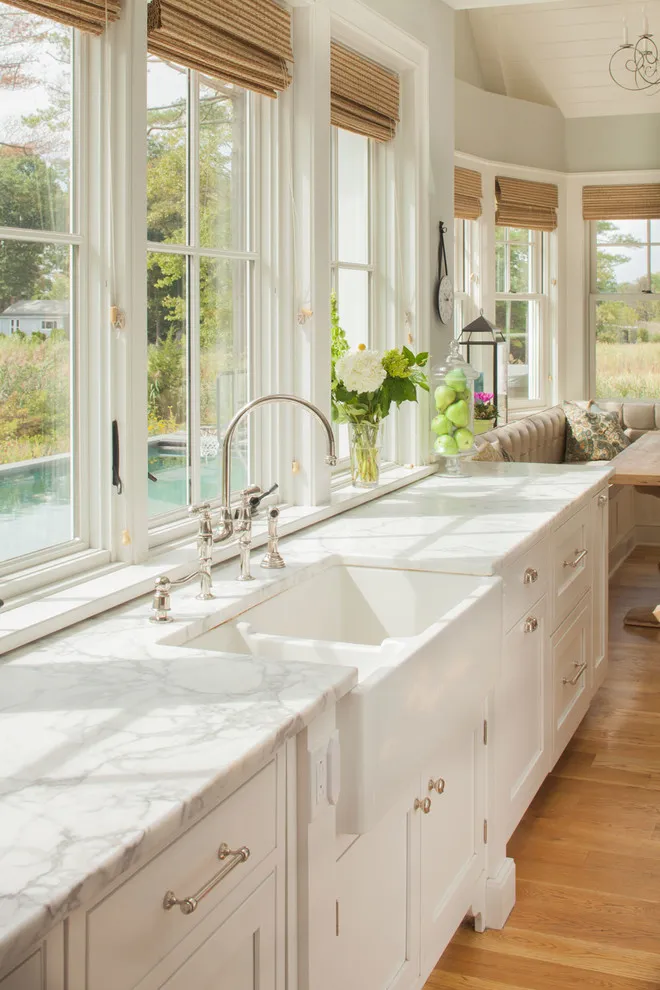 Pale cream cabinets and veined counters run below tall windows with woven shades
