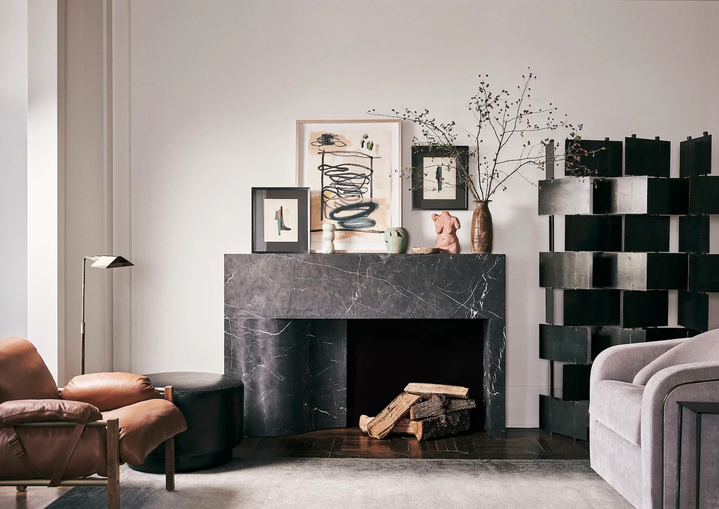 A dark veined rectangular fireplace surround stands beside open black shelving, pale seating and a smaller group of framed pictures and vessels