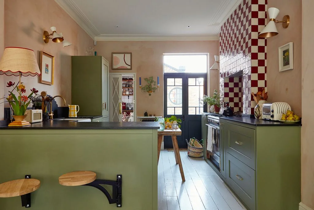 Red-and-white checked tiles rise above green kitchen cabinets beside pink walls, framed pictures and a decorative floral lamp on the counter