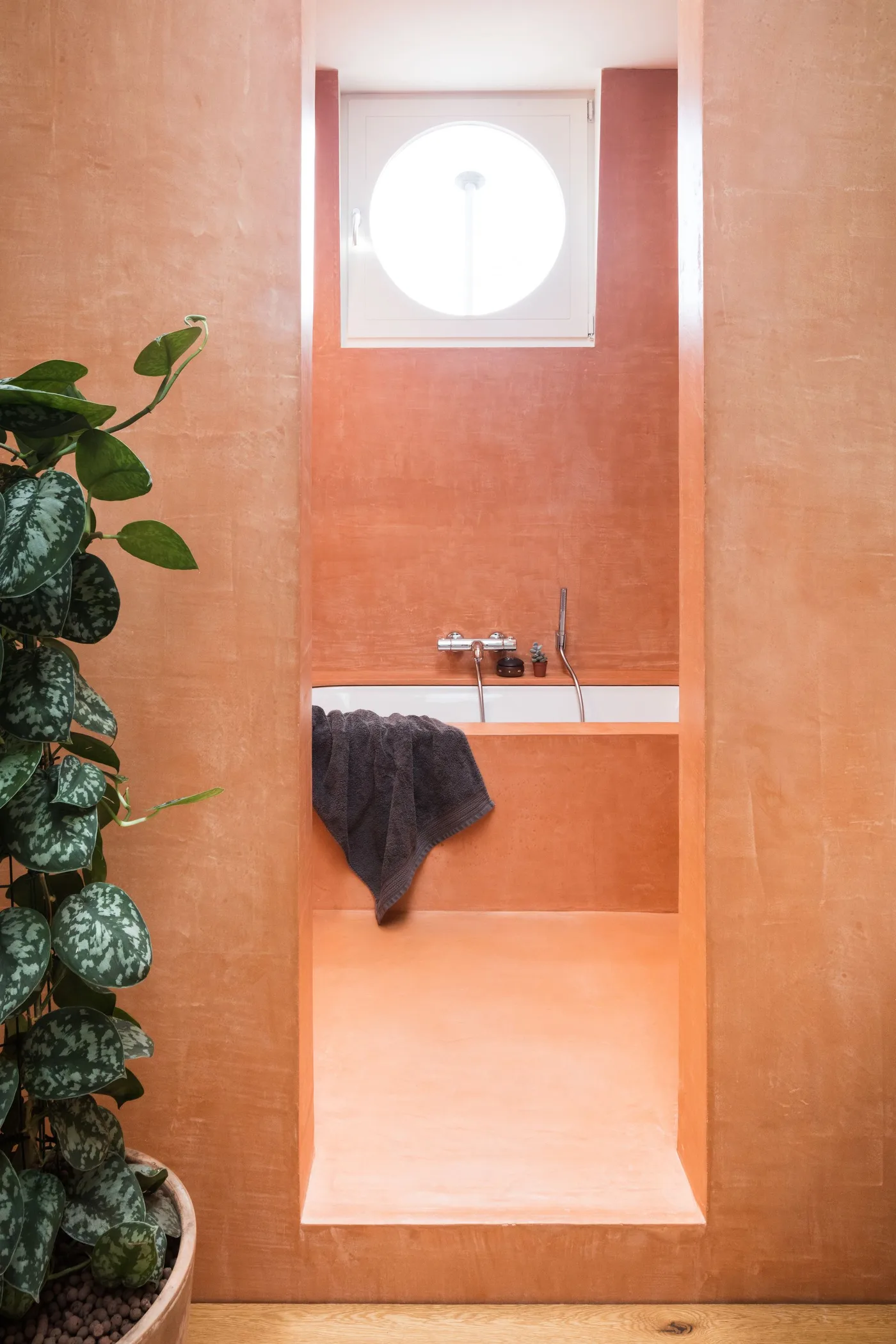 Warm clay-colored bath recess with a circular window, pale inset bath and leafy plant near the opening