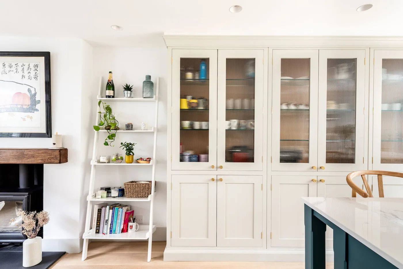 Cottage kitchen with pale glazed cupboard doors, visible wooden shelves, a narrow open shelf unit and the edge of a dark green island.