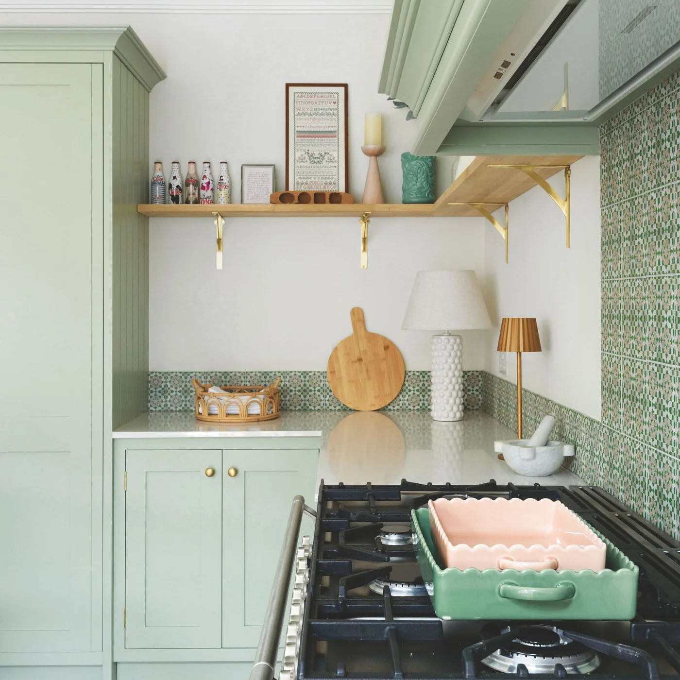 Pale mint green cupboards sit beside green patterned tiles and a pale counter near a cooking surface and wooden shelf