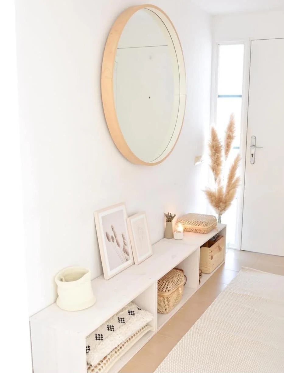 A long pale open-compartment unit holds woven baskets beneath a round wooden mirror beside a white front door and pale rug
