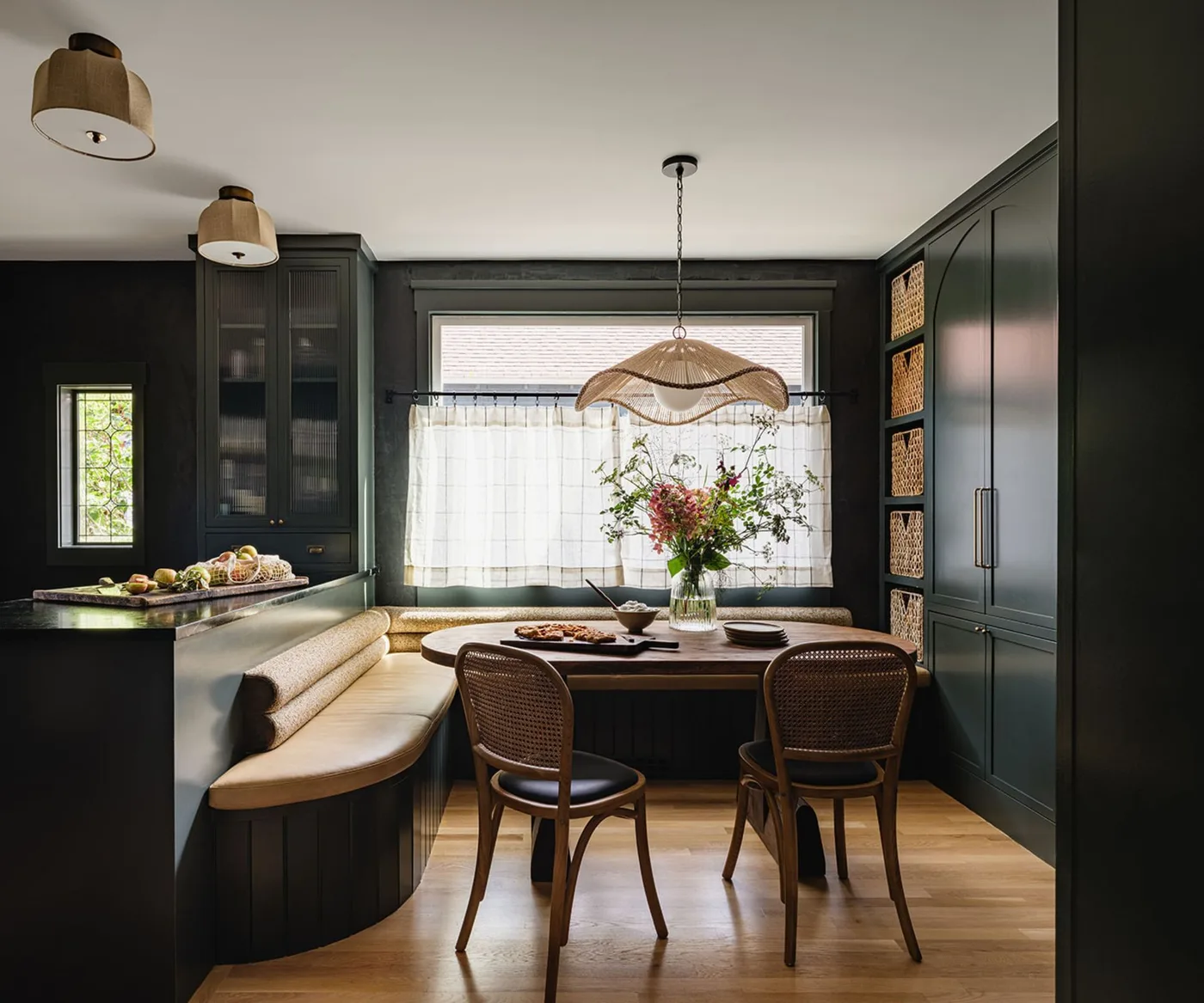 Cottage kitchen dining nook with deep green cabinetry, pale cushioned corner seating, a round wooden table and two curved dark chairs.