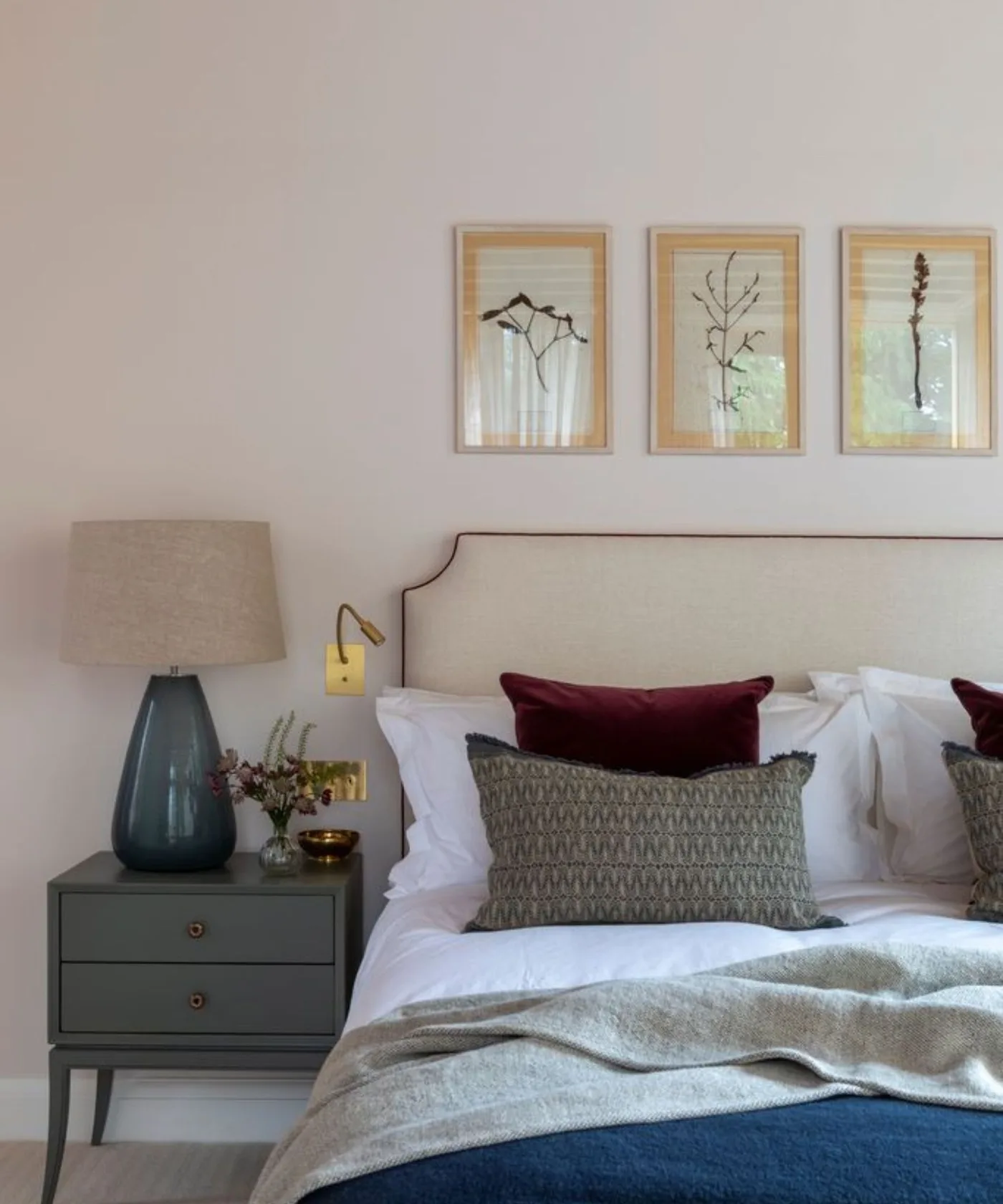 Bedroom with pale shaped headboard, white bedding, darker pillows and a gray bedside cabinet carrying a teal lamp beside a small wall reading light.